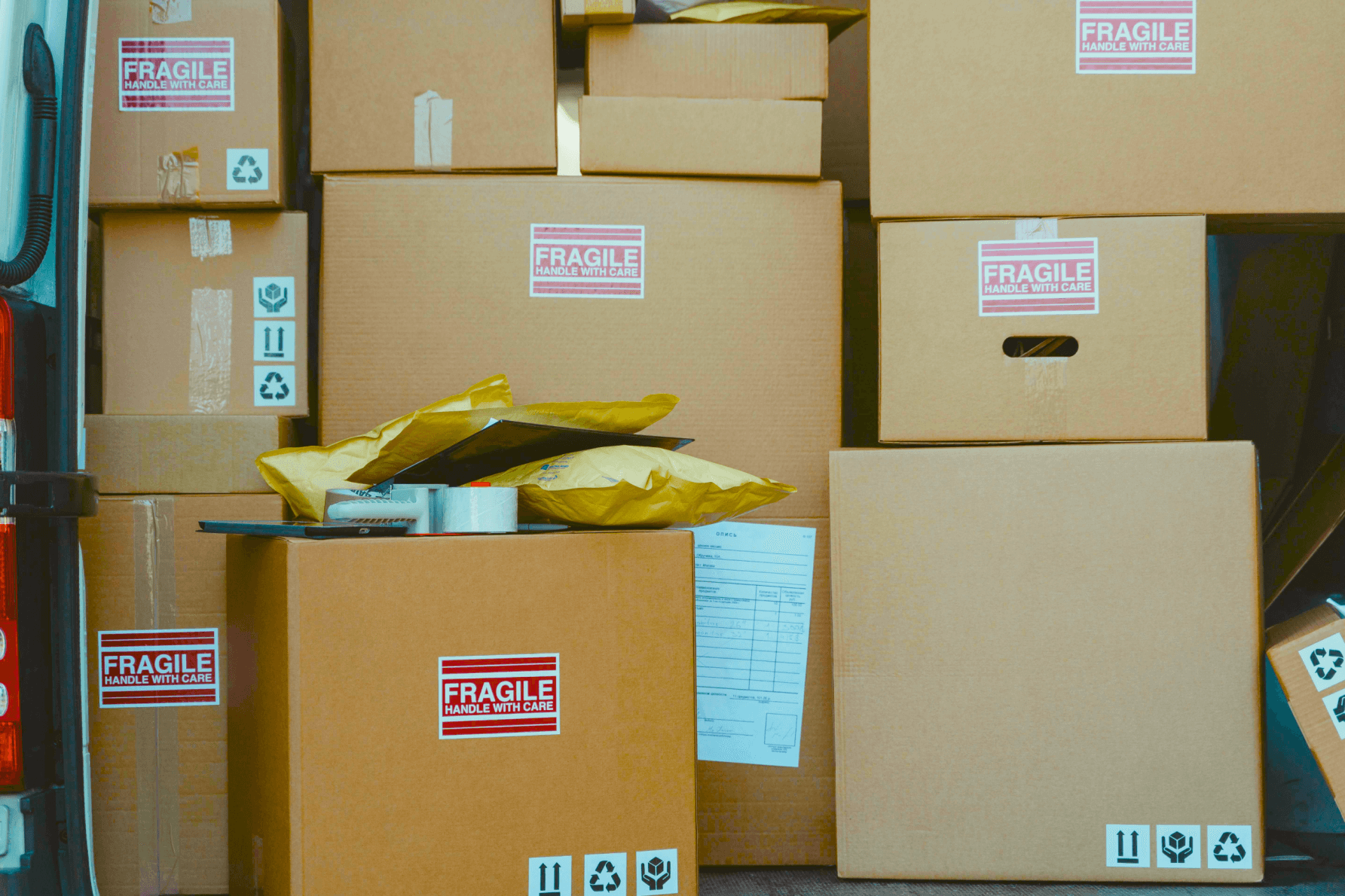  young woman in a yellow shirt is sitting at a desk, talking on the phone and smiling, surrounded by cardboard boxes. The background shows shelves with more packages, indicating a small business or home office setting.