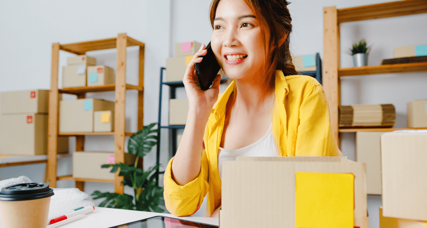  young woman in a yellow shirt is sitting at a desk, talking on the phone and smiling, surrounded by cardboard boxes. The background shows shelves with more packages, indicating a small business or home office setting.